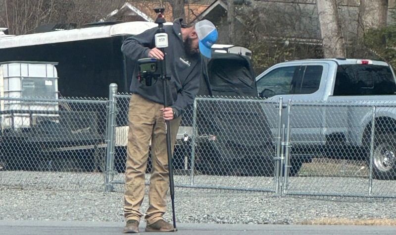 Surveyor using an AutoPole with prism on a survey pole during construction staking beside a gravel roadway.