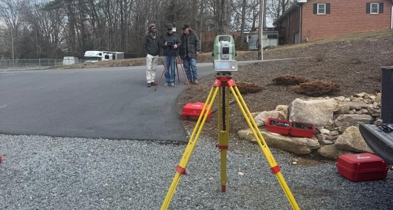 Robotic total station set up on a tripod beside a paved roadway, with a small group of surveyors reviewing data near field equipment in the background