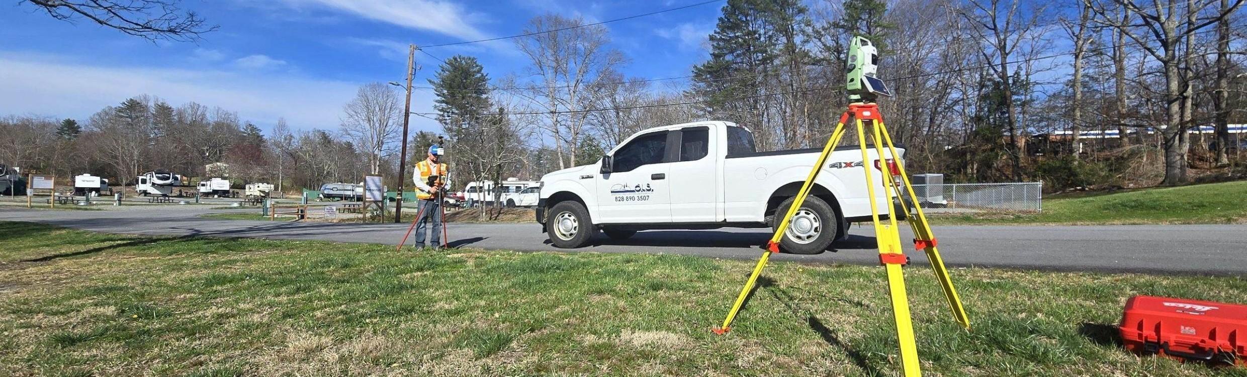 Survey equipment set up beside a roadside, with a robotic total station on a tripod in the foreground and a surveyor collecting measurements near a work vehicle.