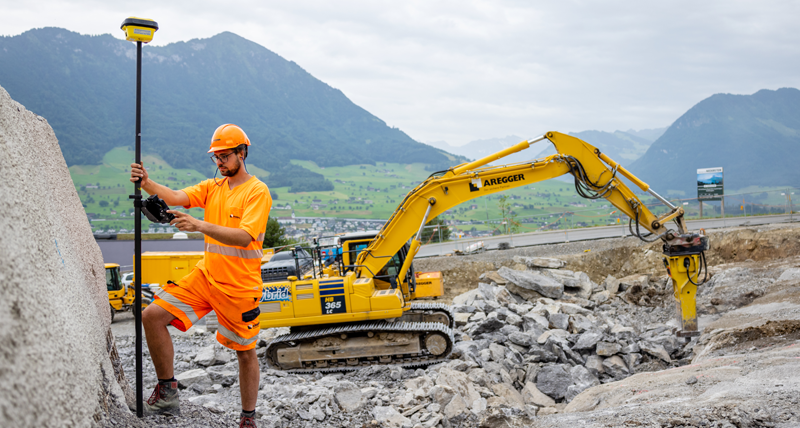 Construction worker in heavy construction measuring with a GNSS antenna and an excavator on the background. 