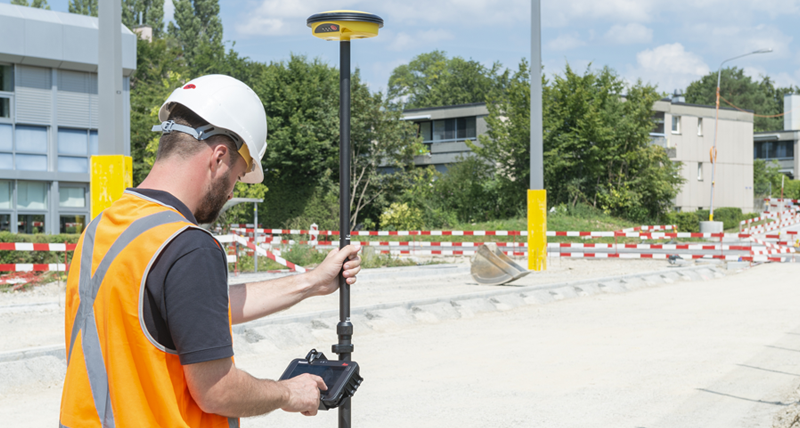 Un ouvrier de chantier portant un gilet de sécurité orange et un casque mesure des points avec un contrôleur de terrain Leica iCON CC70 et une Leica iCON GPS 30.