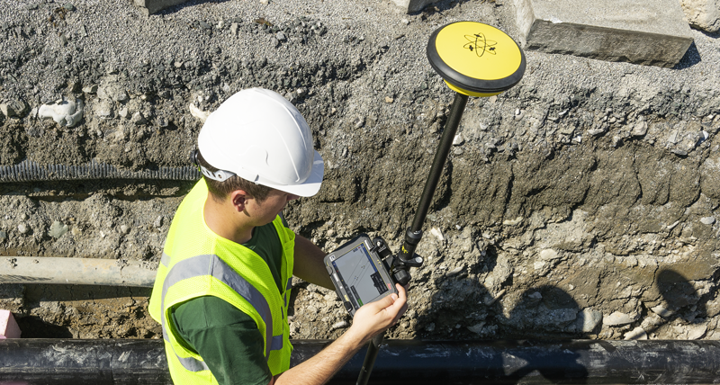 Un ouvrier de chantier portant un gilet de sécurité jaune et un casque dans une tranchée avec des tuyaux enregistre des points avec un carnet de terrain Leica iCON CC70 et une Leica iCON GPS 30.