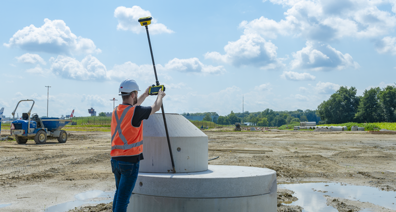 Un ouvrier de chantier portant un gilet de sécurité orange mesure des points dans une zone difficile avec le contrôleur de terrain Leica CC80 et une Leica iCON GPS70.