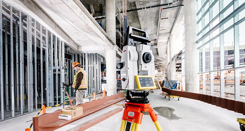 Trabajador de construcción con chaleco naranja y casco en obras de construcción de un edificio, mirando a un controlador de campo Leica iCON CC80 conectado a una estación total robotizada Leica iCON iCR70 para construcción.