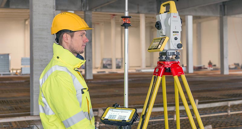 Bauarbeiter in gelber Weste und mit Schutzhelm hält einen Prismenstab mit Feld-Controller Leica iCON CC80 mit Software iCON build, der mit einer Robotic Totalstation Leica iCON iCR80 verbunden ist.