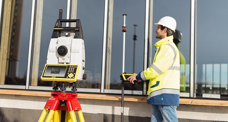 Construction worked in yellow west and hard hat holds a prism pole with the Leica iCON CC80 field controller with iCON build software and is connected to a Leica iCON iCR80 robotic total station with stable target lock during layout.